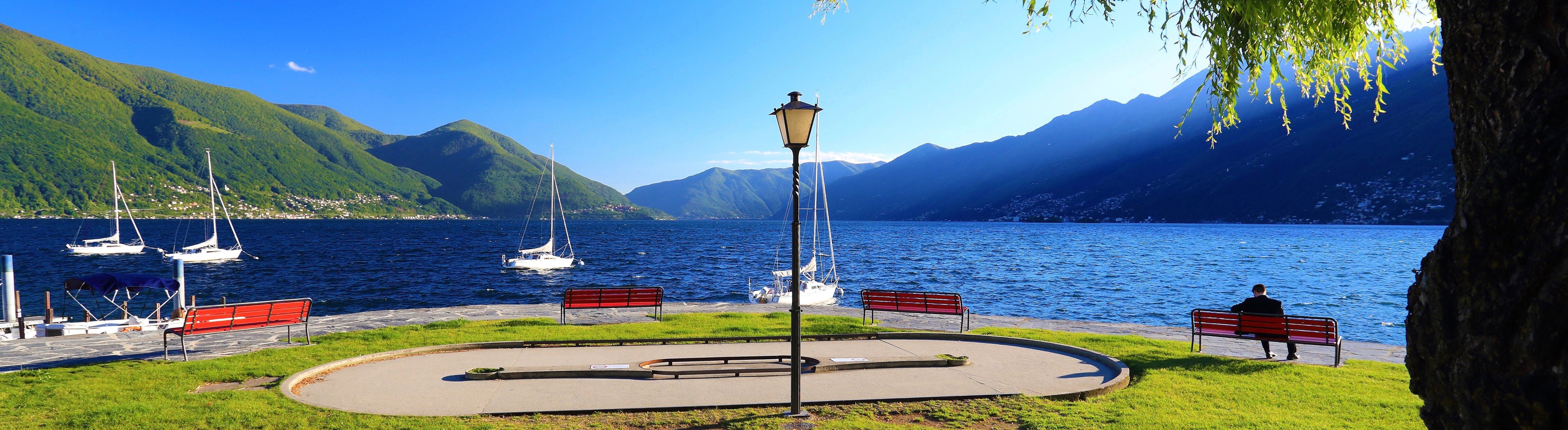 Lakeside square with benches, sailboats, and mountain views across Lago Maggiore